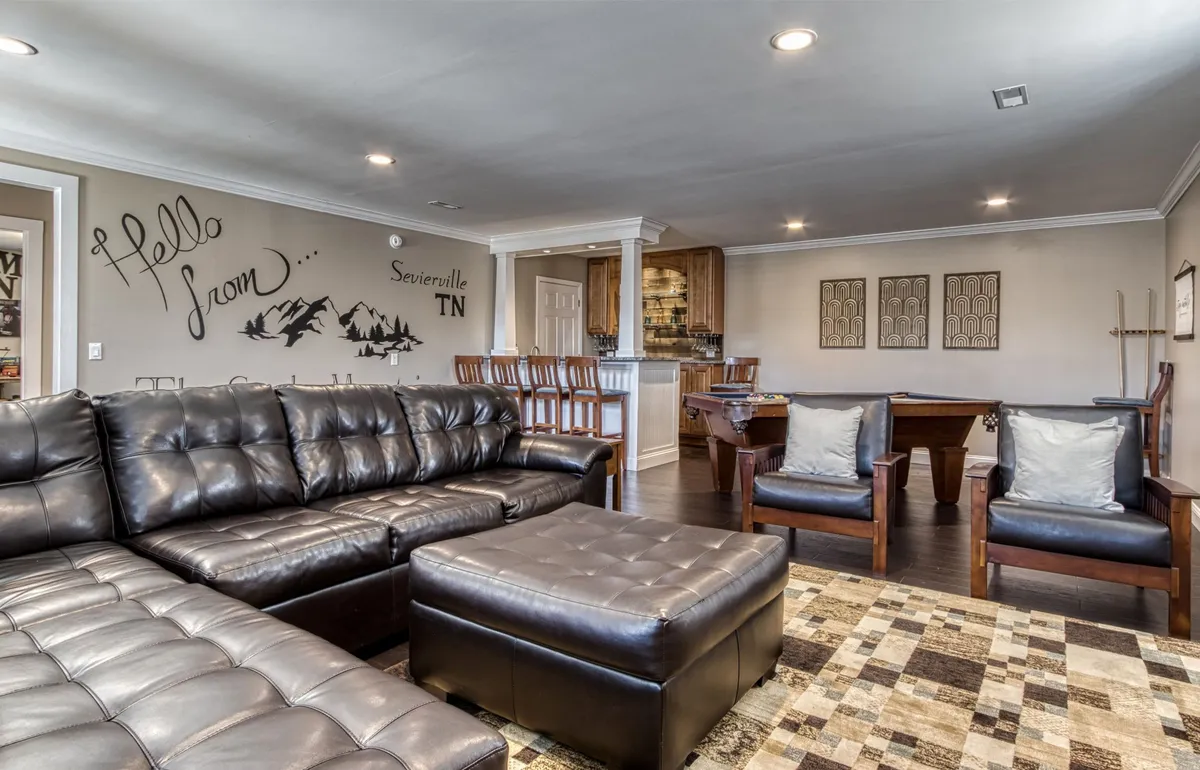 A lively game room setting that includes a leather sectional sofa, a pool table, and a bar area. The space is designed for socializing, friendly competition, and relaxation, with warm lighting and a casual, welcoming atmosphere.