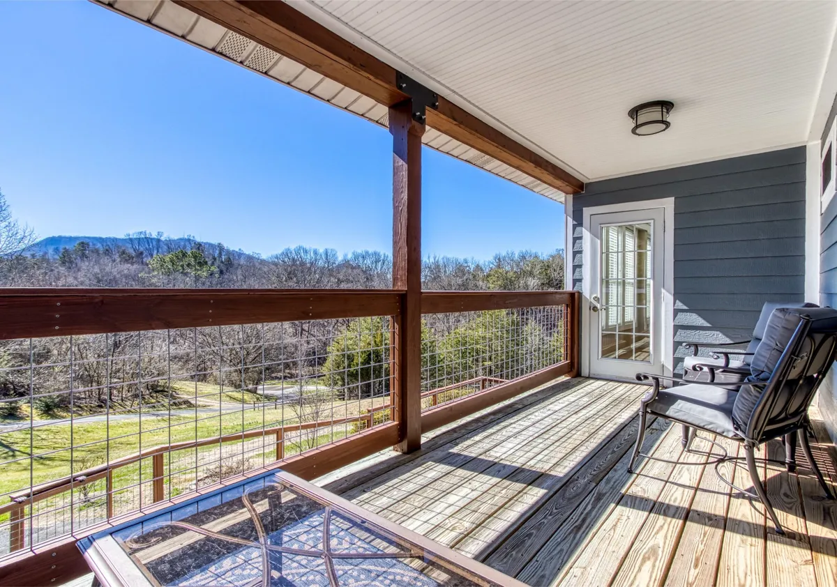 A spacious deck furnished with a glass table and chairs overlooks a sweeping view of the Smoky Mountains. The scene suggests a serene setting ideal for morning coffee, reading, or simply soaking in the quiet beauty of nature. The wooden deck and mountain backdrop reinforce the connection to resort-style outdoor living.