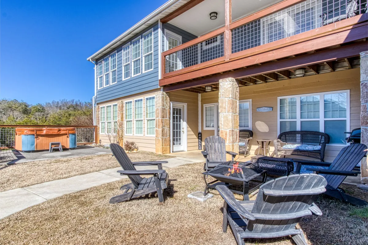 This image beautifully captures the exterior deck area with both the hot tub and fire pit in view, alongside comfortable seating and the exterior of the house. The space is set up for relaxation and social gatherings, framed by the scenic backdrop of the Smoky Mountains. The lighting suggests sunset, adding to the tranquil and inviting atmosphere.