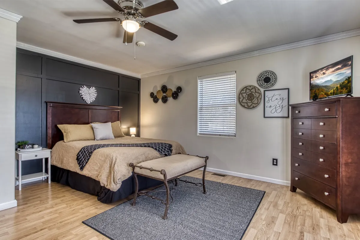A calm bedroom featuring a well-made bed, a nightstand, dresser, wall décor, and a window that lets in gentle natural light. The décor and furnishings emphasize comfort and serenity, with a subtle nod to mountain themes, ideal for couples or families seeking quiet privacy.