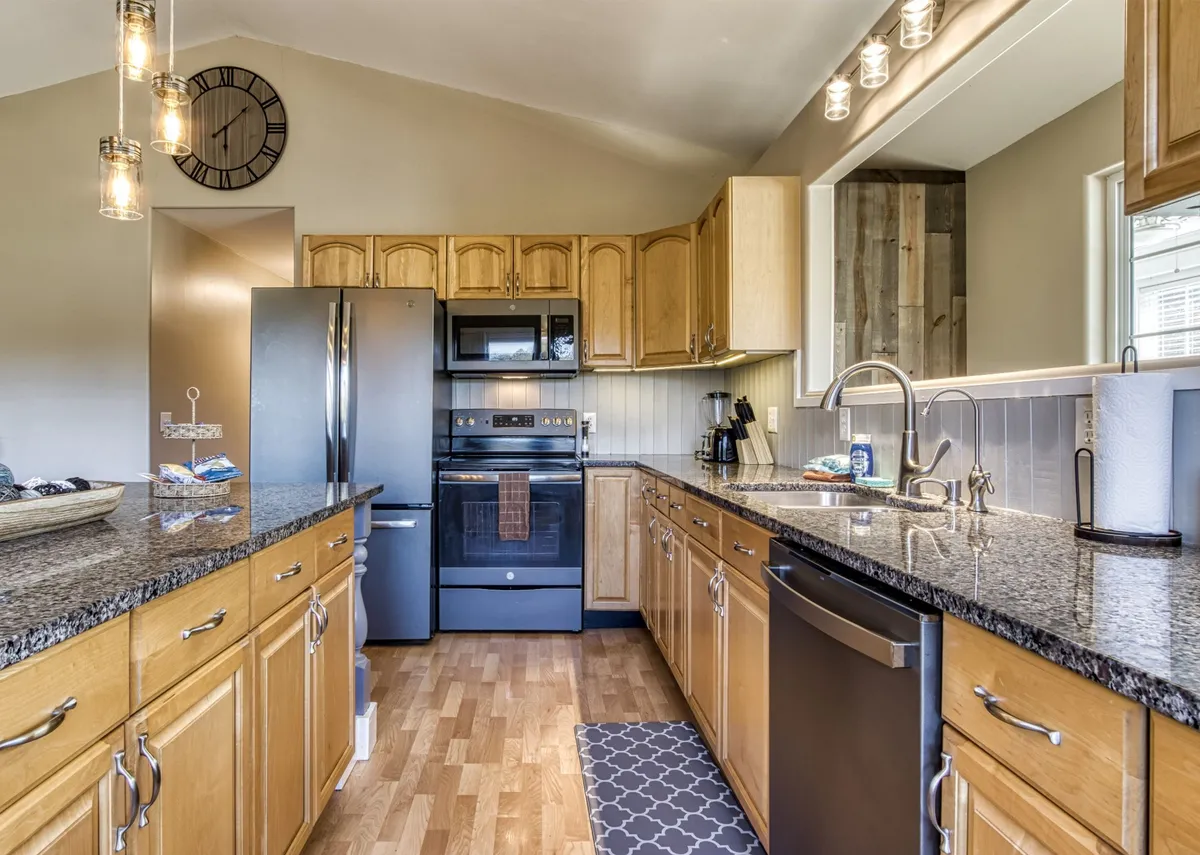 This image features a beautifully designed kitchen with granite countertops, stainless steel appliances, wood cabinetry, a kitchen island, and a decorative clock. The space is open, bright, and clearly set up for meal preparation and group gatherings.