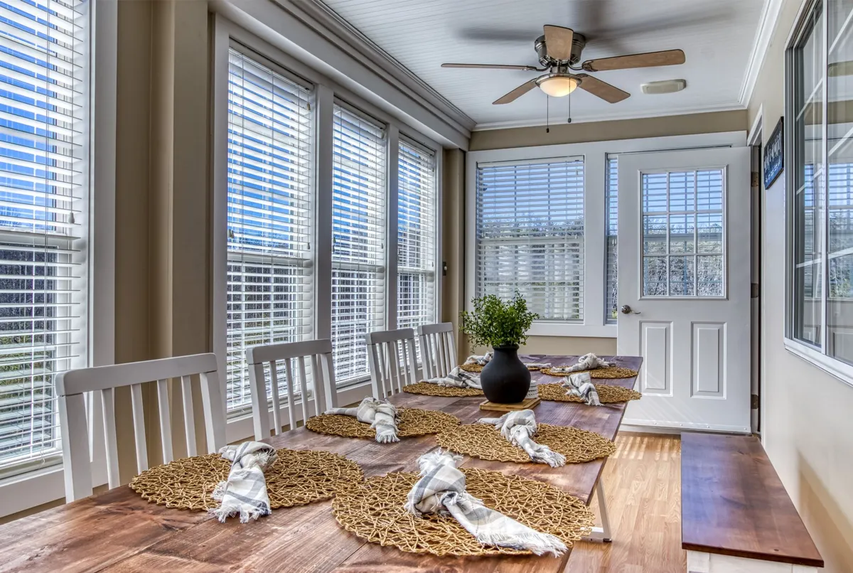 The image shows a warm and inviting dining area with a large dining table, a vase with a plant as centerpiece, and a window with soft natural light. The atmosphere is cozy, ideal for family meals and social gatherings.
