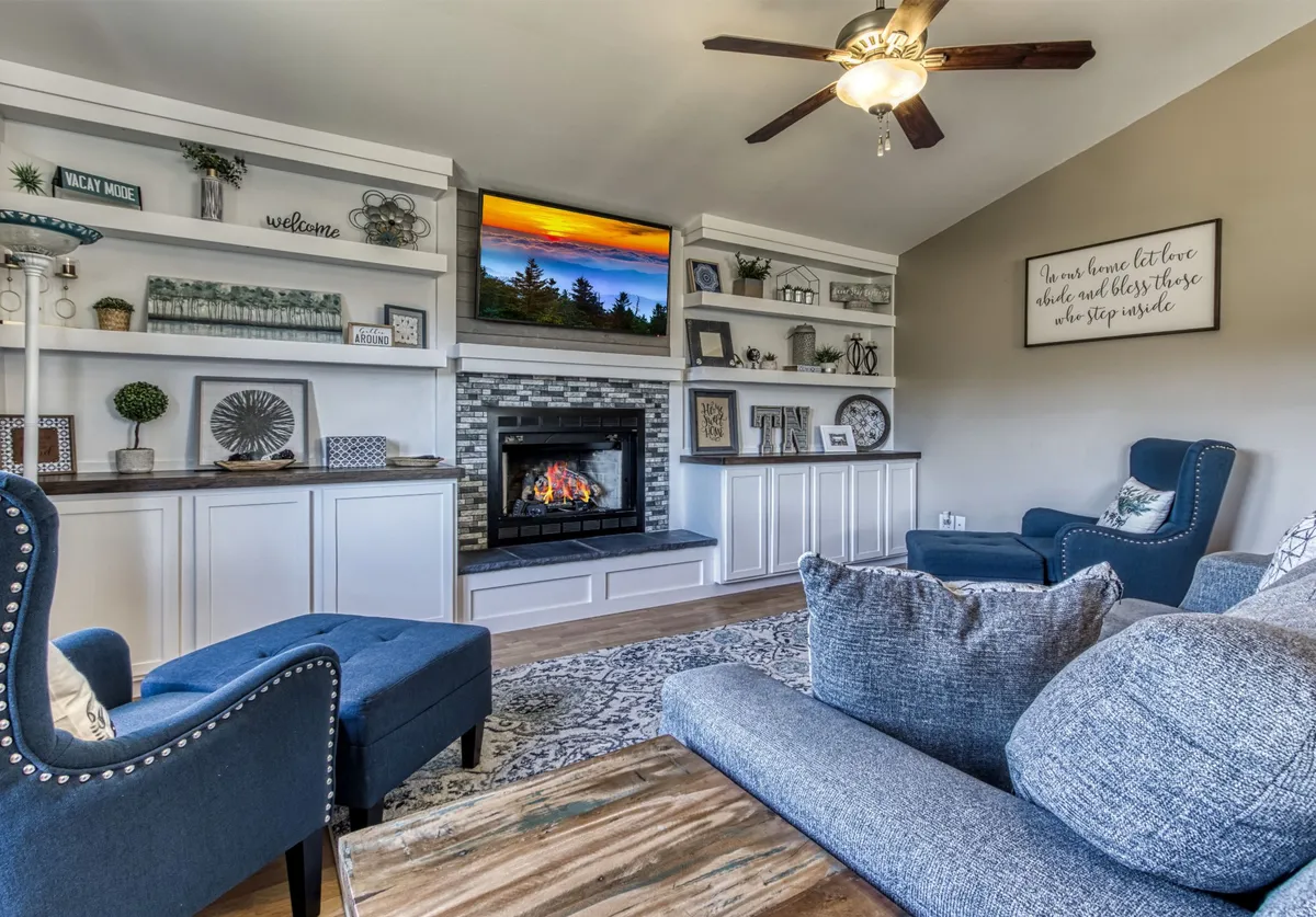 A warm and inviting living room featuring a prominent stone fireplace, shelves adorned with tasteful decor, a comfortable sofa, and a television. The room is styled in cozy, rustic tones, creating the perfect spot for relaxing or gathering with family and friends.