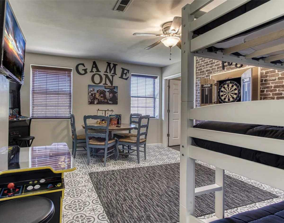A playful interior featuring a bunk bed, an arcade machine, a dartboard, a dining table, and a wall with the phrase 'GAME ON.' The area is vibrant and energetic, ideal for board games, video games, and family fun.